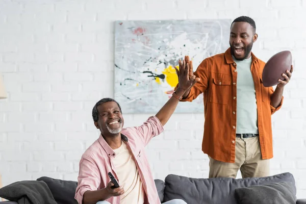 Père et fils afro-américains positifs donnant cinq tout en regardant le match de rugby à la maison — Photo de stock