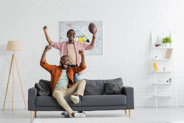 Волнующий африканский отец и сын смотрят матч по регби в современной гостиной — стоковое фото