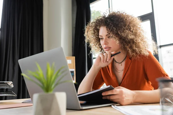 Fokussierte Geschäftsfrau mit Stift und Mappe beim Blick auf Laptop im Büro — Stockfoto