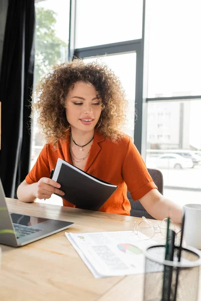 Довольная деловая женщина с кудрявыми волосами, держащая папку возле ноутбука на рабочем столе — стоковое фото