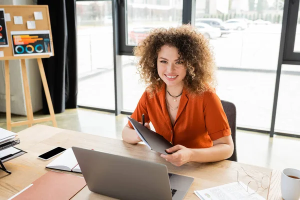Fröhliche Geschäftsfrau mit lockigem Haar hält Ordner in der Nähe von Laptop und Infografiken auf Büroschreibtisch — Stockfoto