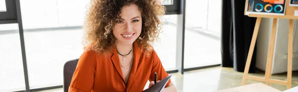 Cheerful businesswoman with curly hair holding folder in modern office, banner — Stock Photo