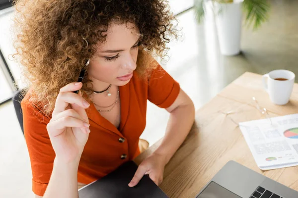 Над головой вид задумчивой деловой женщины, держащей ручку и папку рядом с ноутбуком — стоковое фото