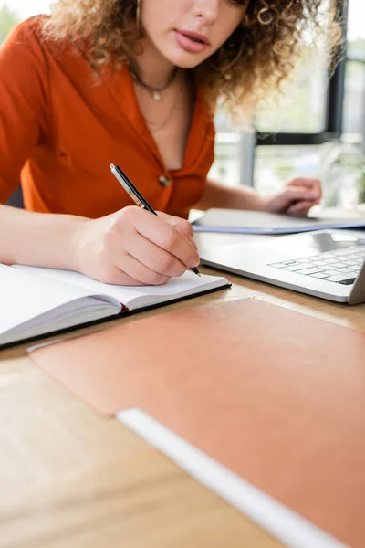 Обрізаний вид фігурної бізнес-леді, роблячи нотатки в ноутбуці біля ноутбука на столі — стокове фото