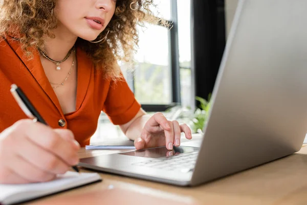 Частичный взгляд на кудрявую деловую женщину, делающую заметки в ноутбуке во время использования ноутбука — стоковое фото