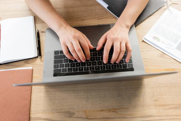 Vista superior da mulher cortada digitando no teclado do laptop perto de pastas e infográficos na mesa — Fotografia de Stock