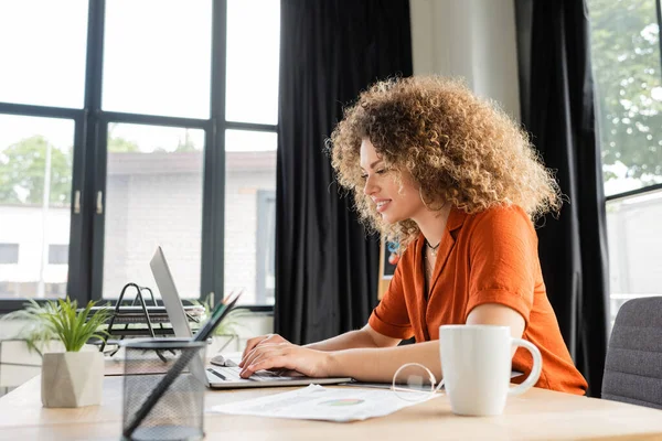 Fröhliche und lockige Geschäftsfrau tippt auf Laptop-Tastatur im modernen Büro — Stockfoto
