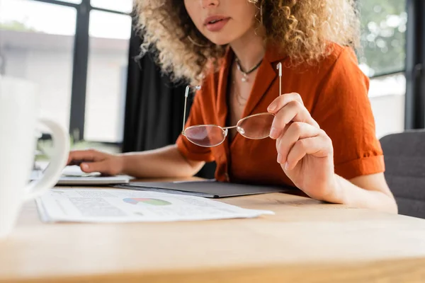 Cropped view of curly woman holding glasses near laptop and infographics in modern office — Stock Photo