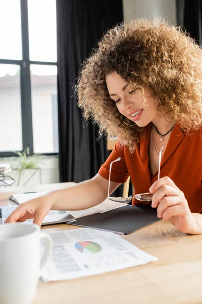 Радостная деловая женщина, держащая очки и глядя на инфографику возле ноутбука и чашку чая — стоковое фото