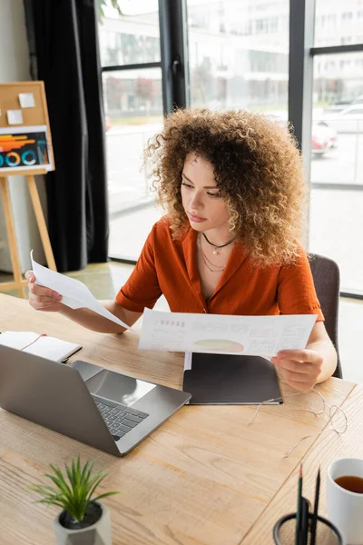 Кудрявая деловая женщина смотрит на инфографику возле ноутбука и чашку чая на столе — стоковое фото