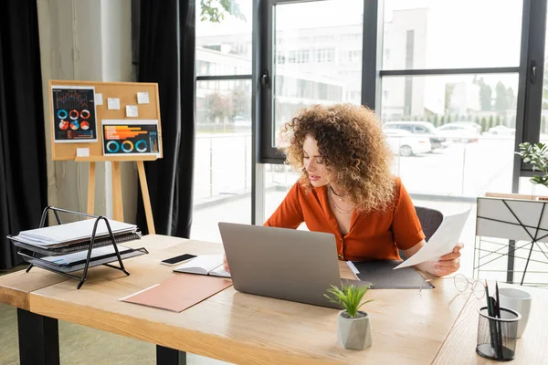 Curly businesswoman holding infographics and using laptop and cup of tea on desk — Stock Photo