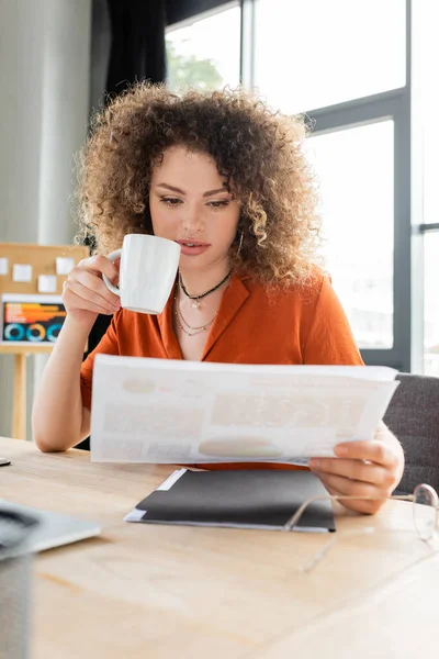 Junge Geschäftsfrau mit lockigem Haar hält eine Tasse Tee in der Hand und betrachtet die Infografiken — Stockfoto