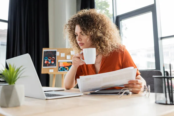 Young businesswoman with curly hair holding cup of tea and infographics while looking at laptop — Stock Photo