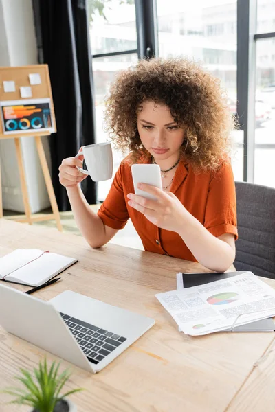 Молода бізнес-леді з кучерявим волоссям тримає чашку чаю і дивиться на смартфон біля ноутбука на столі — стокове фото