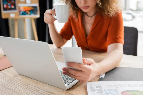 Cropped view of businesswoman holding cup of tea and using smartphone near laptop on desk — Stock Photo