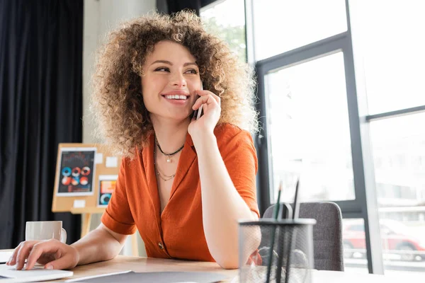 Счастливая деловая женщина разговаривает на смартфоне и с помощью ноутбука возле чашки на столе — стоковое фото