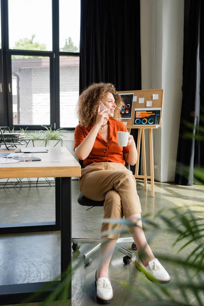 Comprimento total de mulher de negócios feliz falando no smartphone e segurando xícara de chá no escritório — Fotografia de Stock