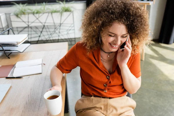 Вид сверху на радостную деловую женщину разговаривающую на смартфоне и держащую чашку чая в офисе — стоковое фото