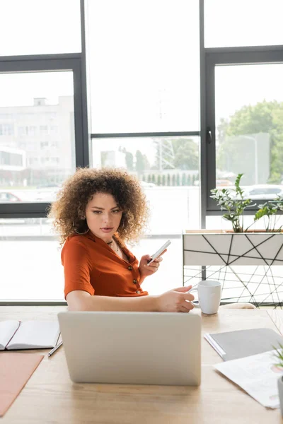 Lockige Geschäftsfrau hält Smartphone und Tasse Tee in der Hand, während sie im Büro auf Laptop schaut — Stockfoto