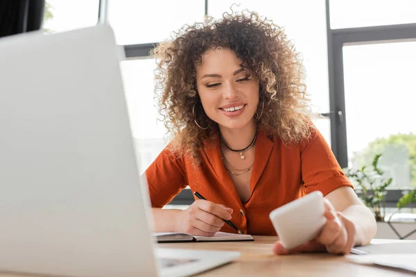 Счастливая деловая женщина, пишущая в ноутбуке и глядя на смартфон рядом с ноутбуком — стоковое фото