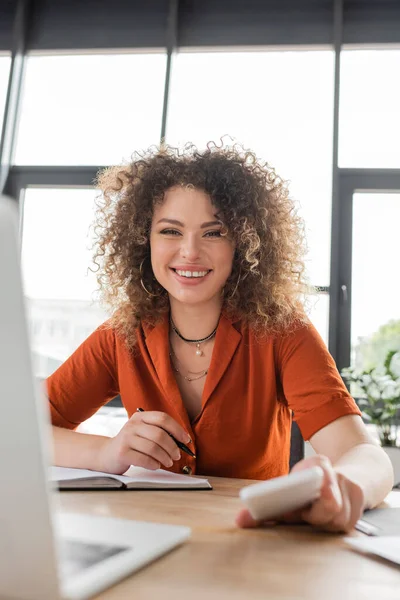 Mulher de negócios feliz com cabelo encaracolado olhando para a câmera e segurando smartphone perto de laptop borrado na mesa — Fotografia de Stock