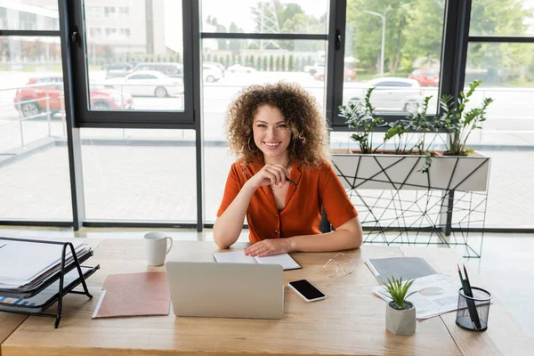 Zufriedene Geschäftsfrau mit lockigem Haar blickt in die Kamera in der Nähe von Gadgets und Dokumenten auf dem Schreibtisch — Stockfoto