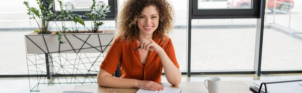 Pleased businesswoman with curly hair looking at camera and holding pen in office, banner — Stock Photo