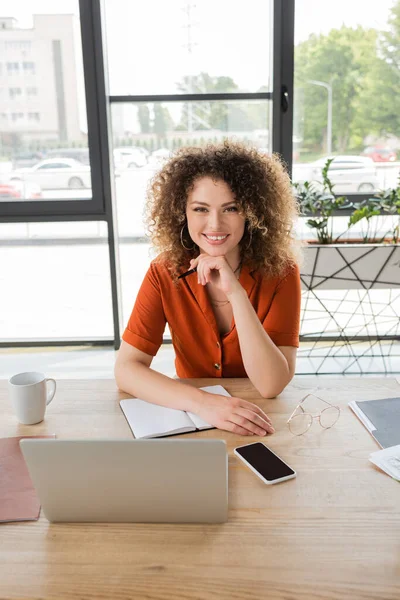Радостная деловая женщина с вьющимися волосами, смотрящая в камеру рядом с гаджетами и чашкой кофе на столе — стоковое фото
