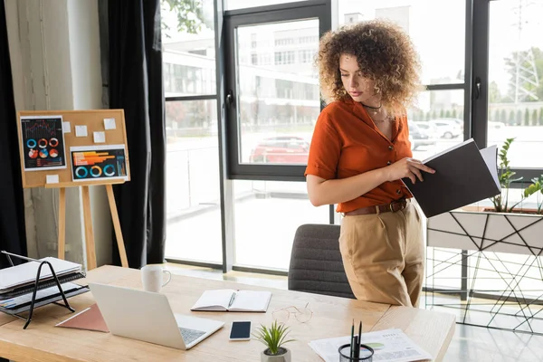 Geschäftsfrau mit lockigem Haar hält Ordner in der Hand und betrachtet Gadgets auf dem Schreibtisch im Büro — Stockfoto