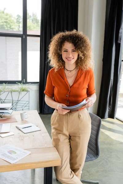 Happy businesswoman with curly hair holding folder and looking at camera in office — Stock Photo