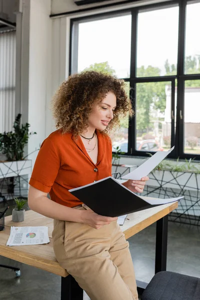 Cheerful businesswoman with curly hair looking at document and holding folder in office — Stock Photo