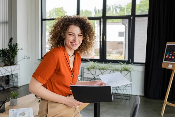 Cheerful businesswoman with curly hair holding folder and paper with graphs in office — Stock Photo