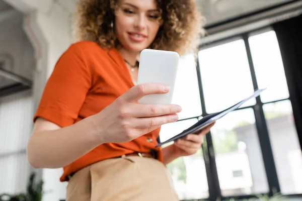 Low angle view of cheerful businesswoman with curly hair holding folder and smartphone in office — Stock Photo