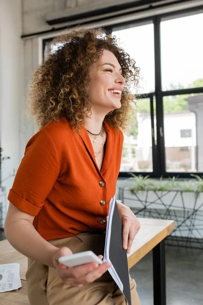 Fröhliche Geschäftsfrau mit lockigem Haar hält Ordner und Smartphone in der Hand, während sie im Büro lacht — Stockfoto