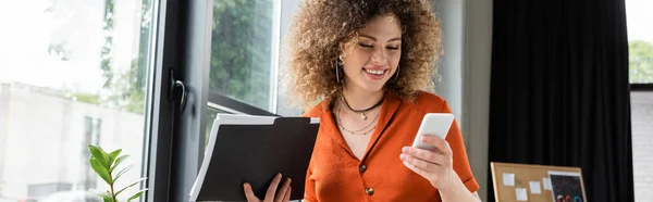 Cheerful businesswoman with curly hair holding folder and using smartphone in office, banner — Stock Photo