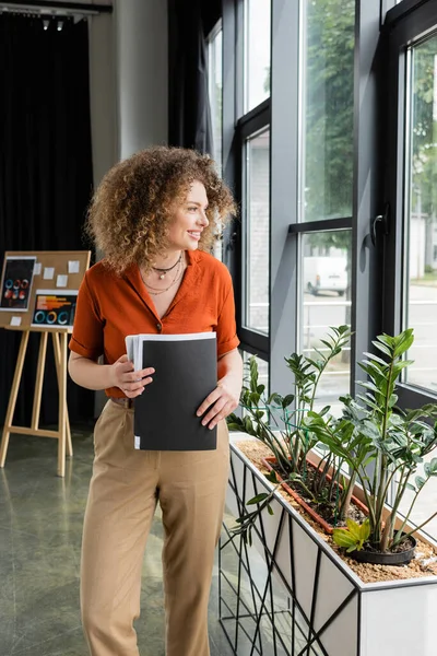 Happy businesswoman with curly hair holding folder and looking at window in modern office — Stock Photo