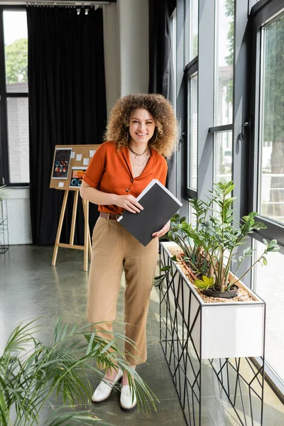 Full length of cheerful businesswoman with curly hair holding folder and looking at camera in modern office — Stock Photo