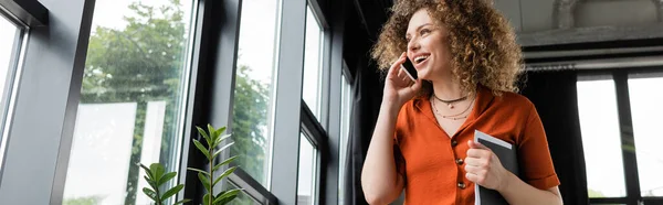 Niedrigwinkel-Ansicht einer glücklichen Geschäftsfrau, die einen Ordner in der Hand hält und im modernen Büro mit dem Smartphone spricht, Banner — Stockfoto