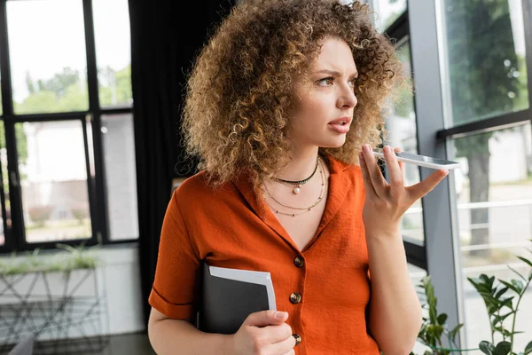 Mulher de negócios séria com cabelo encaracolado segurando pasta e gravação de mensagem de voz no smartphone no escritório moderno — Fotografia de Stock
