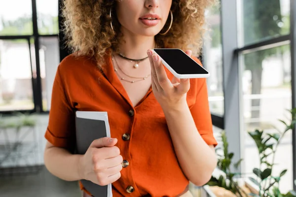 Cropped view of businesswoman with curly hair holding folder and recording voice message on smartphone in office — Stock Photo
