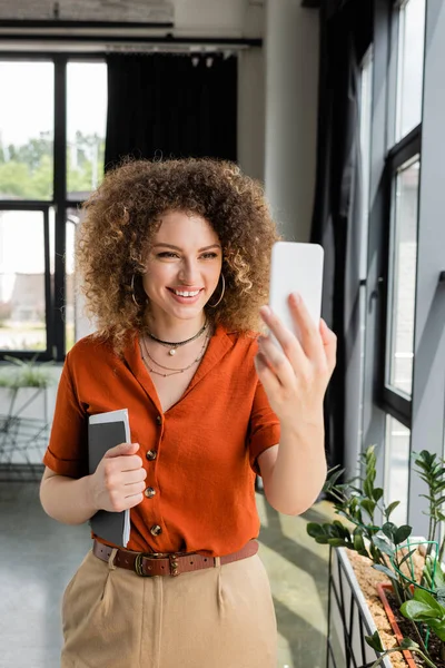 Весела бізнес-леді з кучерявим волоссям, що тримає папку і має відеодзвінок на смартфон в сучасному офісі — стокове фото
