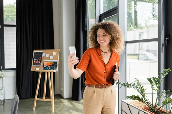 Mujer de negocios complacida con la carpeta de retención de pelo rizado y tener videollamada en el teléfono inteligente en la oficina moderna - foto de stock