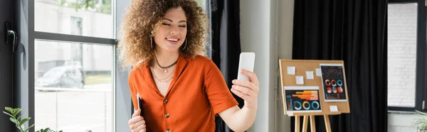 Pleased businesswoman with curly hair holding folder and having video call on smartphone in office, banner — Stock Photo