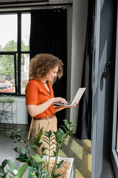 Side view of happy businesswoman with curly hair using laptop next to green plants in modern office — Stock Photo