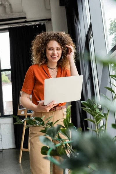 Mulher de negócios positiva com cabelo encaracolado segurando laptop ao lado de plantas verdes no escritório moderno — Fotografia de Stock