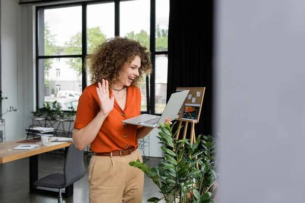 Joyful businesswoman with curly hair having video call and waving hand at laptop in modern office — Stock Photo