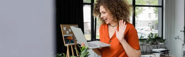 Joyful businesswoman with curly hair having video call and waving hand at laptop in modern office, banner — Stock Photo