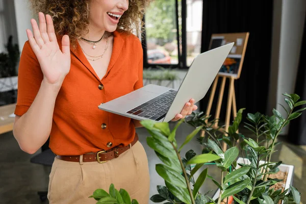 Vista ritagliata di donna d'affari gioiosa con capelli ricci che hanno videochiamata e mano ondulante a computer portatile in ufficio — Foto stock