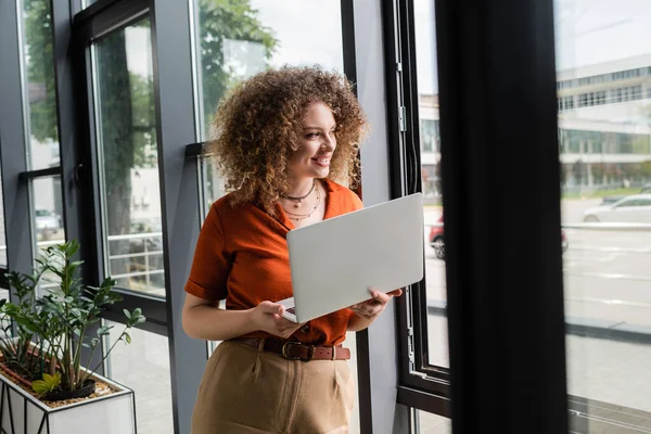 Щаслива бізнес-леді з кучерявим волоссям тримає ноутбук і дивиться через вікно в сучасному офісі — стокове фото