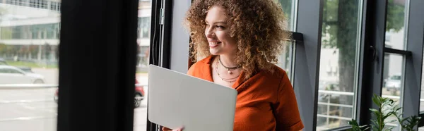 Mulher de negócios feliz com cabelo encaracolado segurando laptop e olhando através da janela no escritório moderno, banner — Fotografia de Stock